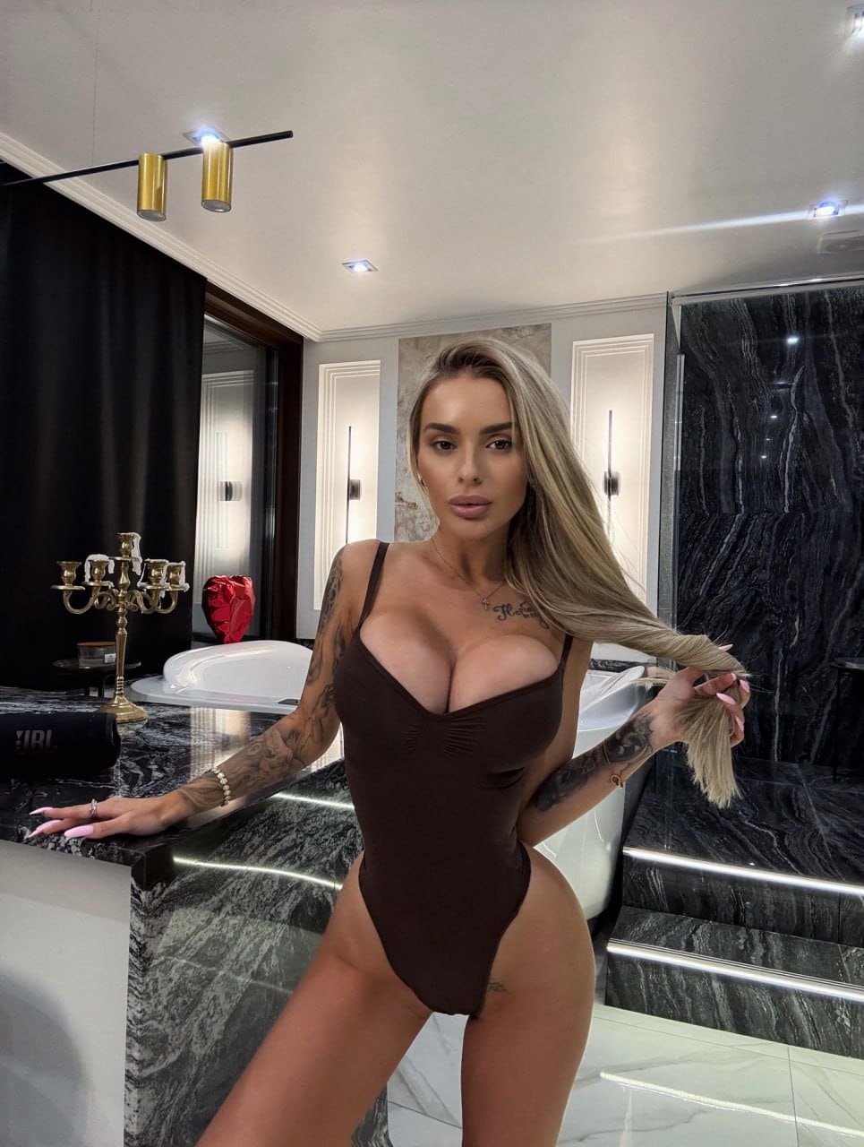 In a marble bathroom, a blonde woman in a deep-plunge brown bodysuit rests one hand on a counter while holding her hair with the other.