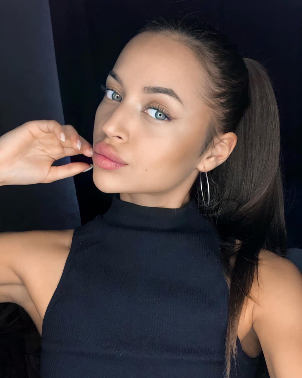 Woman with high ponytail in black turtleneck and hoop earrings poses with hand raised near face against dark background.