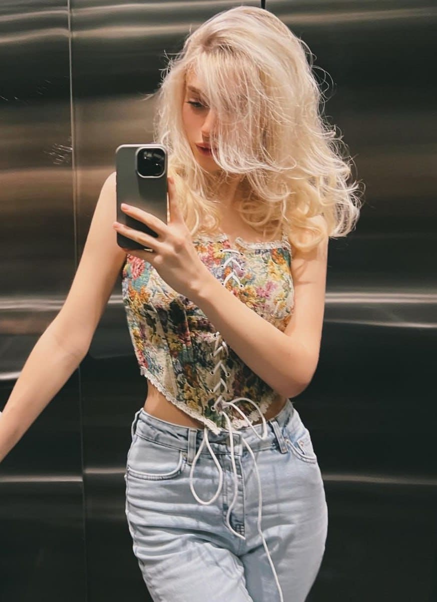 Laura takes a mirror selfie in an elevator, dressed in a floral lace-up corset top with spaghetti straps and light-wash blue jeans.
