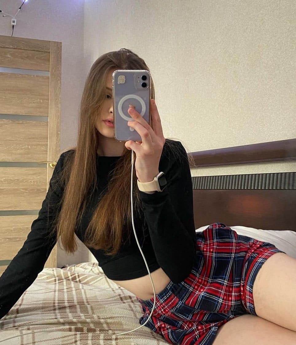 Sitting on a bed, she takes a mirror selfie in a black long-sleeved crop top and a red plaid mini skirt that exposes her legs.