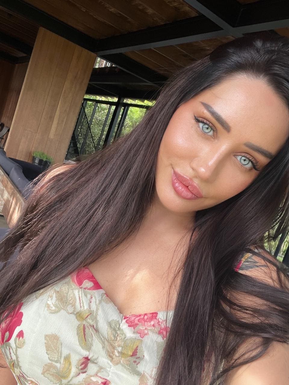 A close-up selfie shows her in a floral sweetheart-neckline top against wooden paneling, with greenery visible in the background.