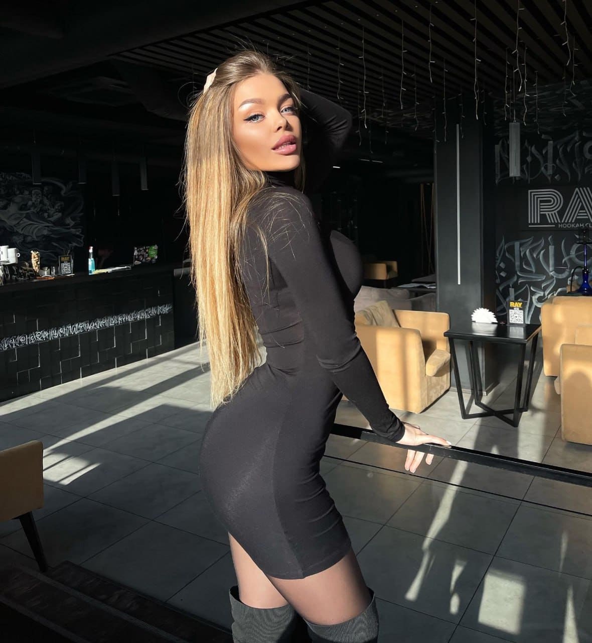 Standing in profile in a lounge, a blonde woman wears a tight black mini dress and thigh-high boots, one hand resting on her head.