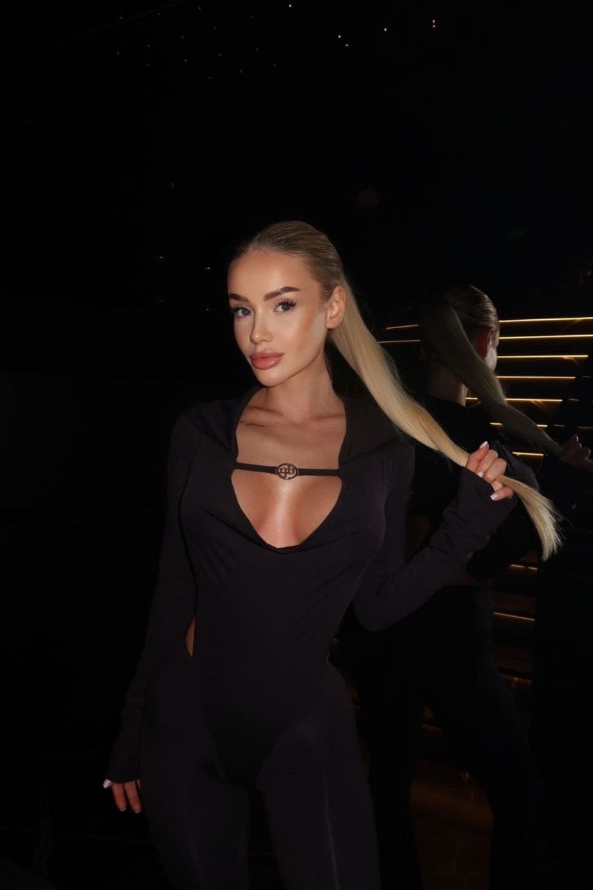 In a dim setting, Belle stands holding her high ponytail, wearing a black long-sleeved bodysuit with a deep neckline and side cutouts.
