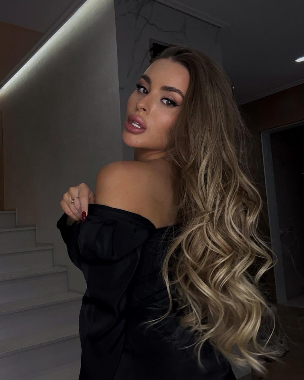 Selena stands in profile over her shoulder in a black off-shoulder top with voluminous sleeves, set against a staircase and marble wall.