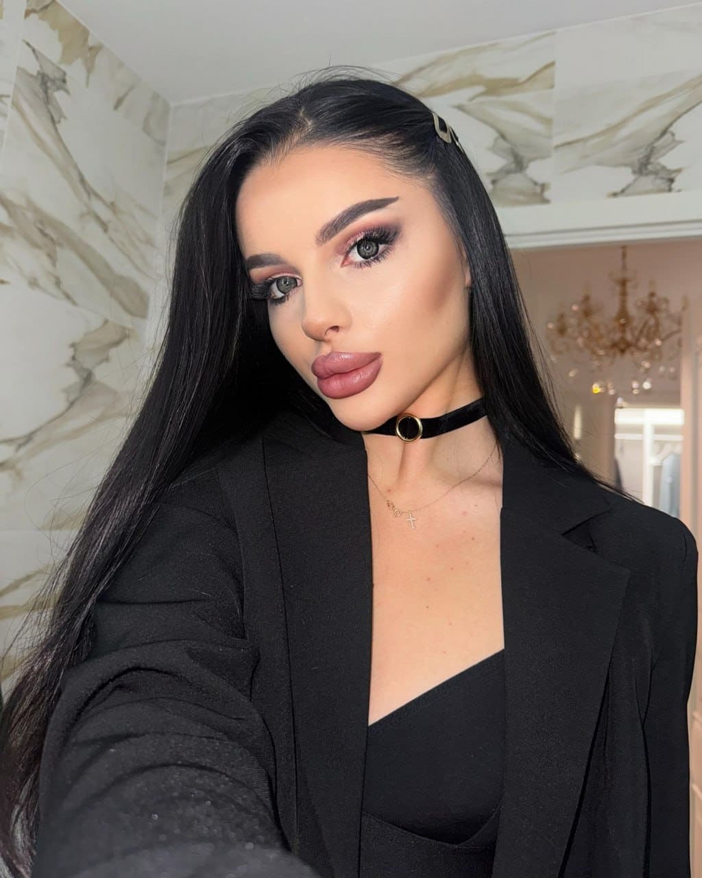 Veronica takes a selfie in a black blazer and top with a choker, posing against a marble-tiled wall with long black hair and a side clip.