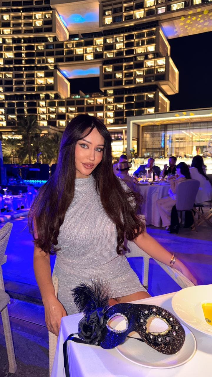 Seated at an outdoor table at night, she wears a short silver sequined dress with a high neckline and a mask on the table.