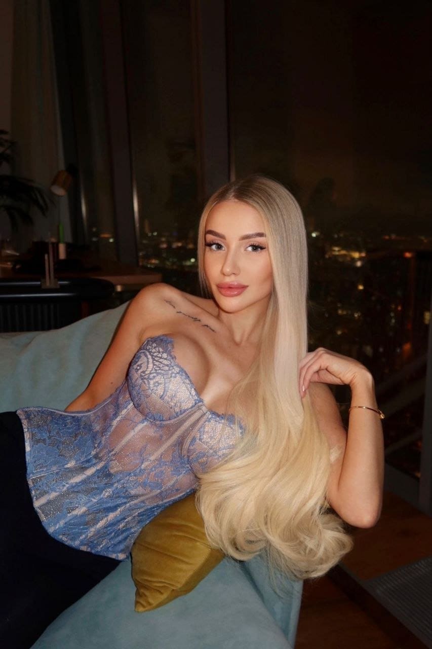 Belle reclines on a light blue sofa in a sheer blue lace corset, with a night city view visible against the dark background.