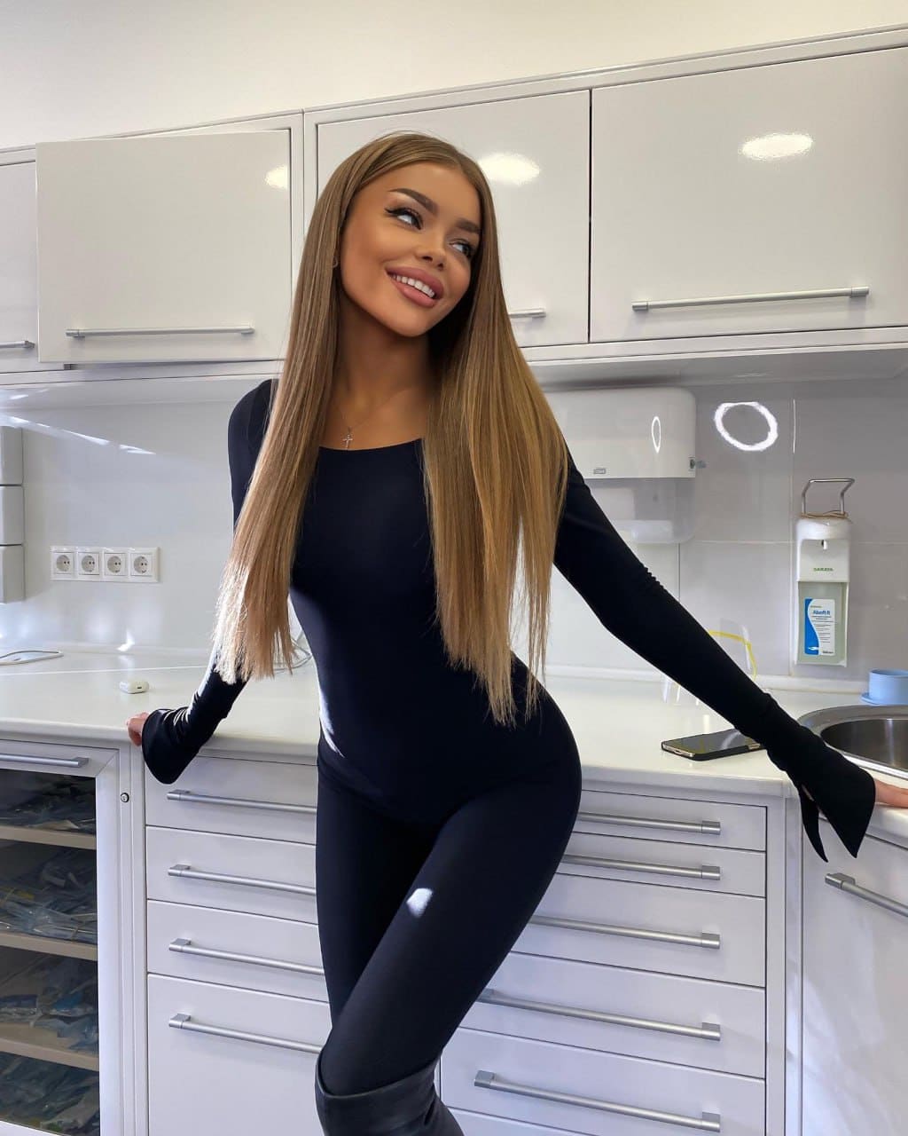 Halia leans against a white kitchen counter in a black long-sleeved bodysuit, leggings, and tall boots, smiling off to the side.
