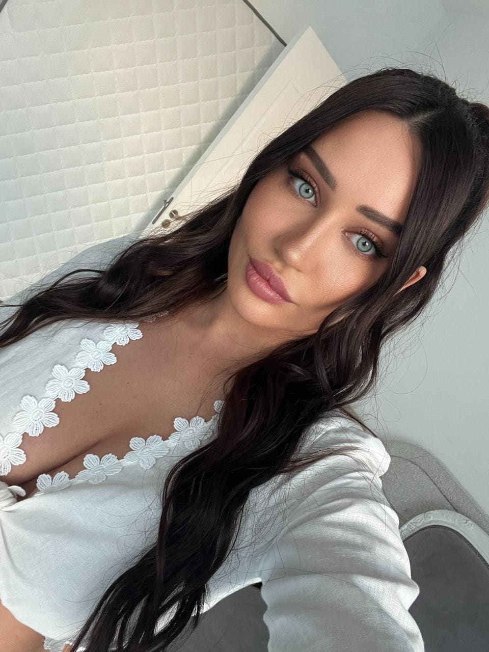 Yasmin takes a selfie in a white plunging-neckline top with lace trim, posed against a white quilted wall and door.