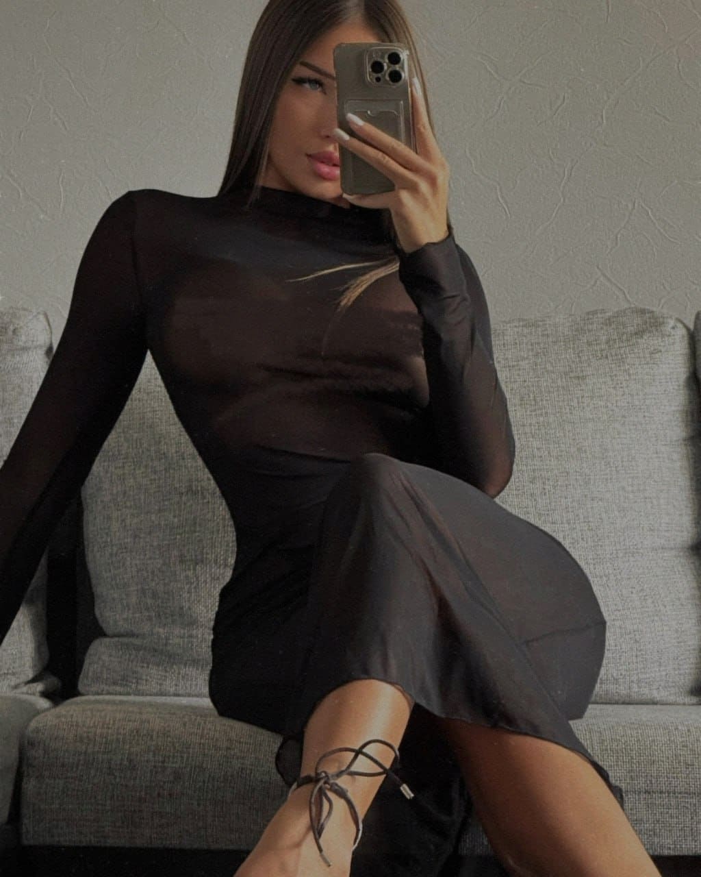 Seated on a gray sofa, Lana takes a mirror selfie in a sheer black midi dress and strappy heels.
