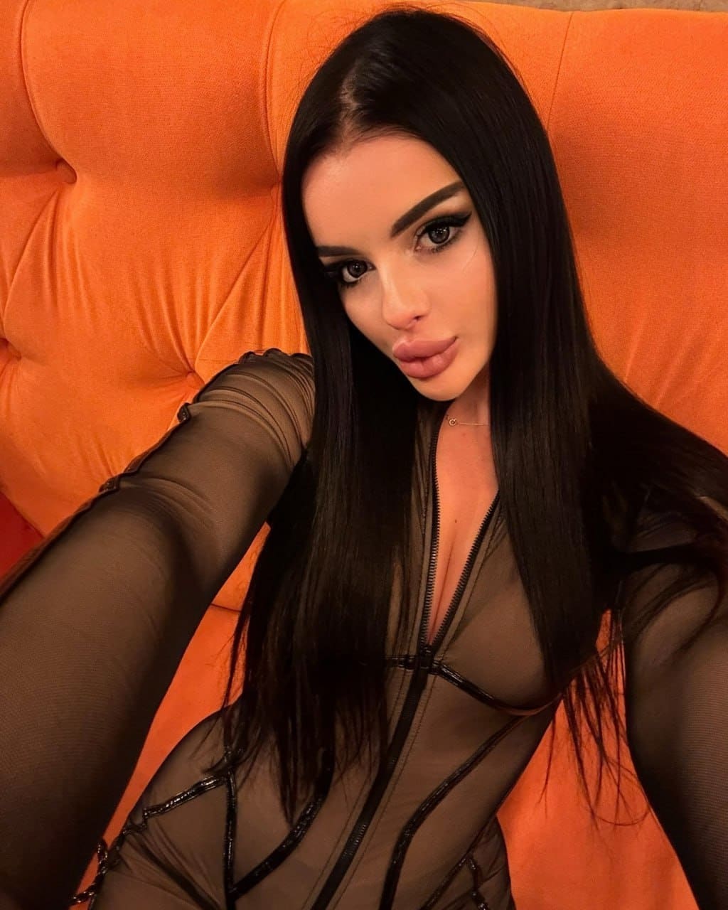 Reclining against an orange tufted headboard, she wears a sheer black bodysuit with opaque piping and a front zipper.