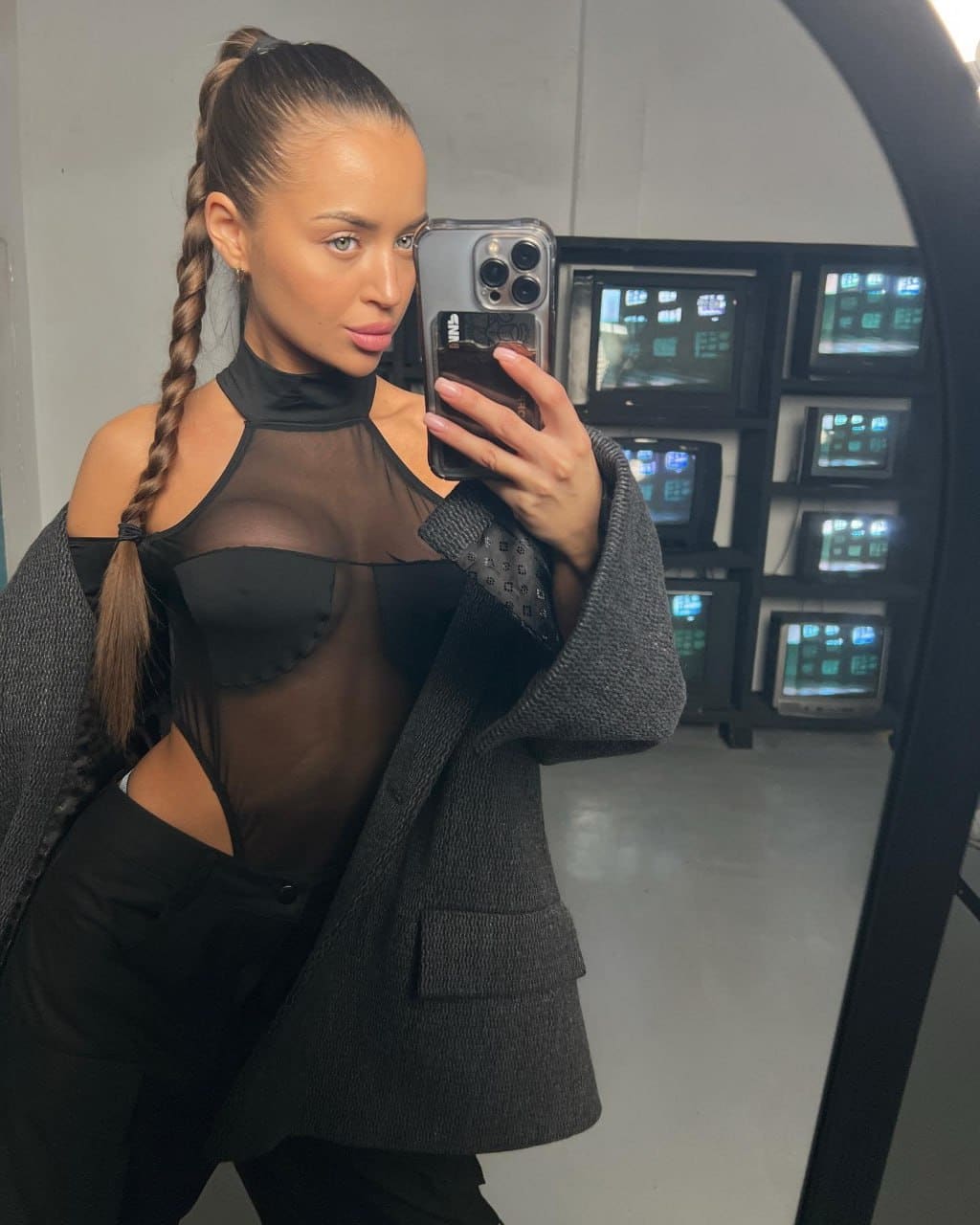 Mirror selfie of a woman with braided ponytail in sheer black bodysuit, trousers, and grey blazer before vintage monitors.