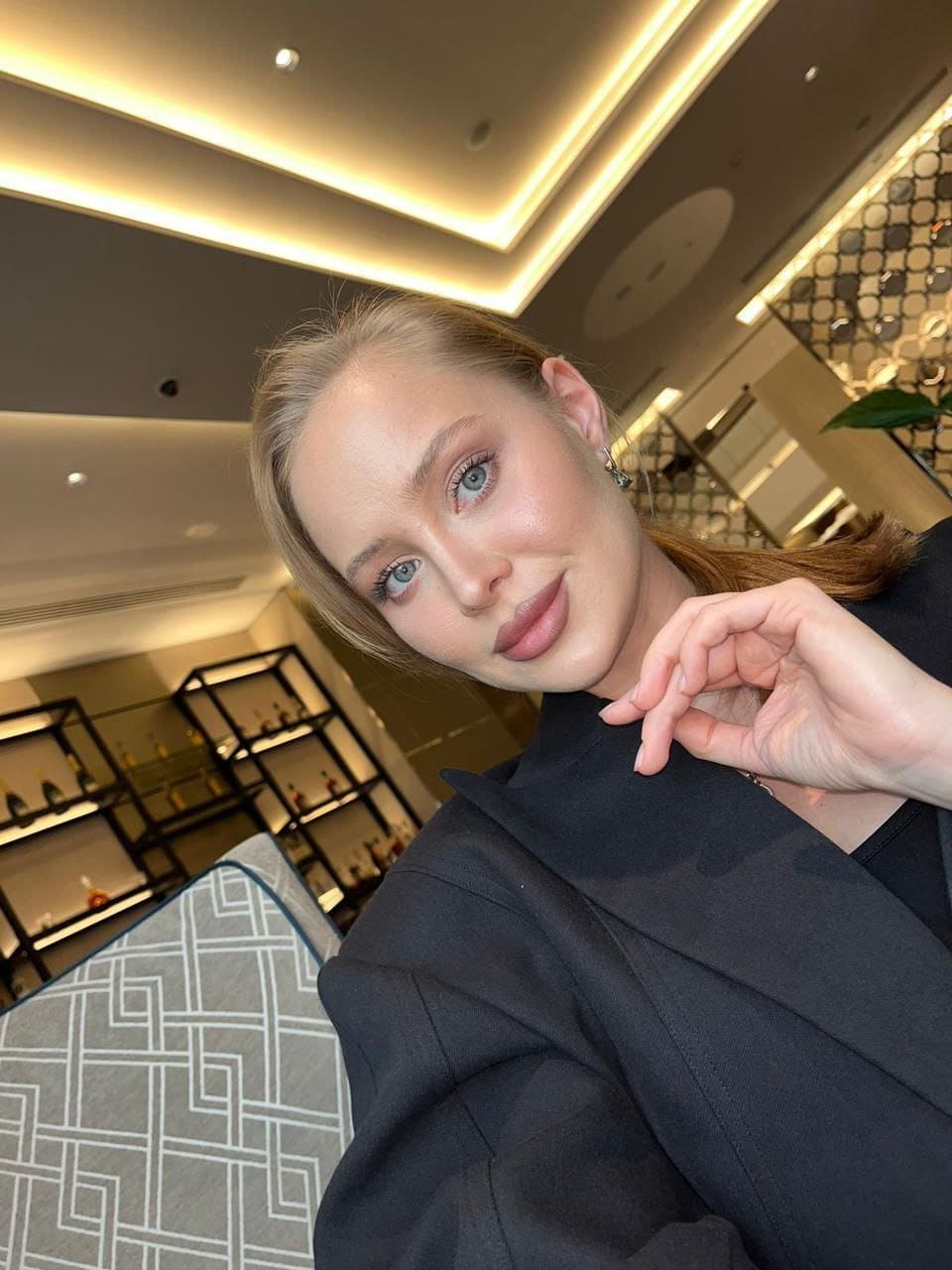 Brooke wears a black blazer and top, posing with a hand near her neck in an indoor setting with warm lighting and shelving.